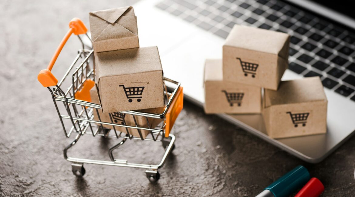 selective focus of toy shopping cart with small carton boxes near laptop, e-commerce concept