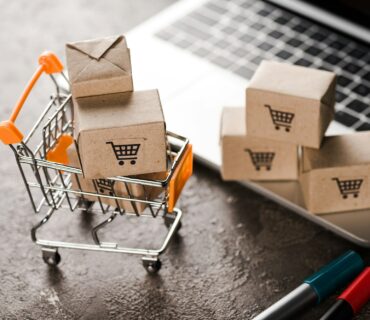 selective focus of toy shopping cart with small carton boxes near laptop, e-commerce concept